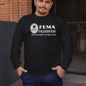 A man wearing a black long-sleeve t-shirt with the text FEMA EVACUATION PLAN RUN MOTHER FUCKER RUN and a Department of Homeland Security seal.
