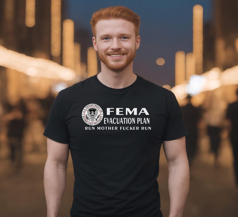 A smiling red-haired man wearing a black crewneck t-shirt featuring a FEMA evacuation plan design.