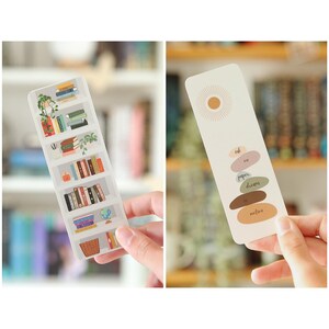 May include: Two bookmarks with different designs. The first bookmark features a bookshelf with books and a globe. The second bookmark has a sun and a stack of colorful stones with the words "soft", "ears", "paper", "dreams", "sun", and "motion" written on them.