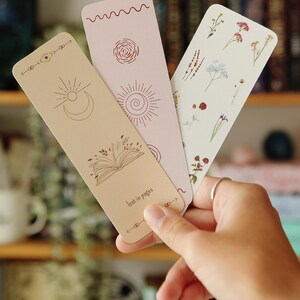 May include: Three bookmarks with different designs. One bookmark features a sun, moon, and open book with the text "lost in pages". Another bookmark has a floral design with pressed flowers. The third bookmark has a sun and a spiral design.