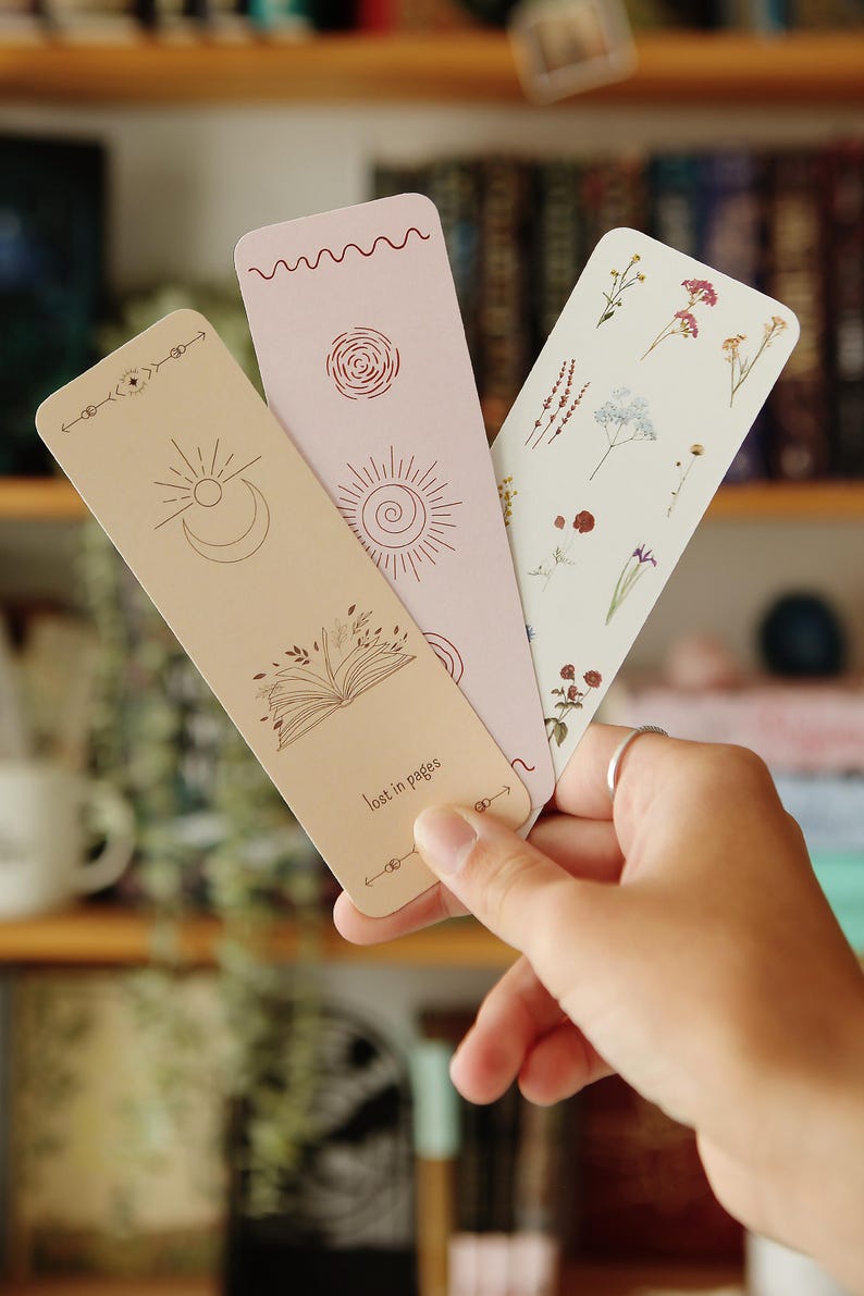 May include: Three bookmarks with different designs. One bookmark features a sun, moon, and open book with the text "lost in pages". Another bookmark has a floral design with pressed flowers. The third bookmark has a sun and a spiral design.