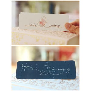 May include: Two bookmark designs. The first bookmark is white with a pink butterfly and the text "ich bin wundervoll". The second bookmark is dark blue with a white crescent moon, stars, and the text "keep dreaming".