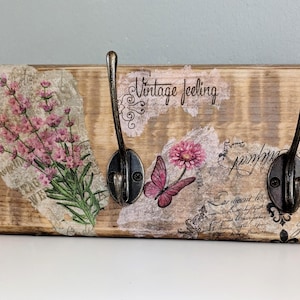 Könnte beinhalten: Wandgarderobe aus Holz mit drei Metallhaken im antiken Stil. Die Garderobe ist mit Blumen- und Schmetterlingsmotiven, Schriftzügen und dem Schriftzug "Vintage feeling" verziert. Die Farben sind Braun, Rosa und Schwarz.