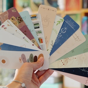 May include: A set of 12 colorful bookmarks with various designs, including floral patterns, celestial motifs, and abstract shapes. The bookmarks are arranged in a fan shape and feature a variety of colors, including pink, blue, green, and brown.