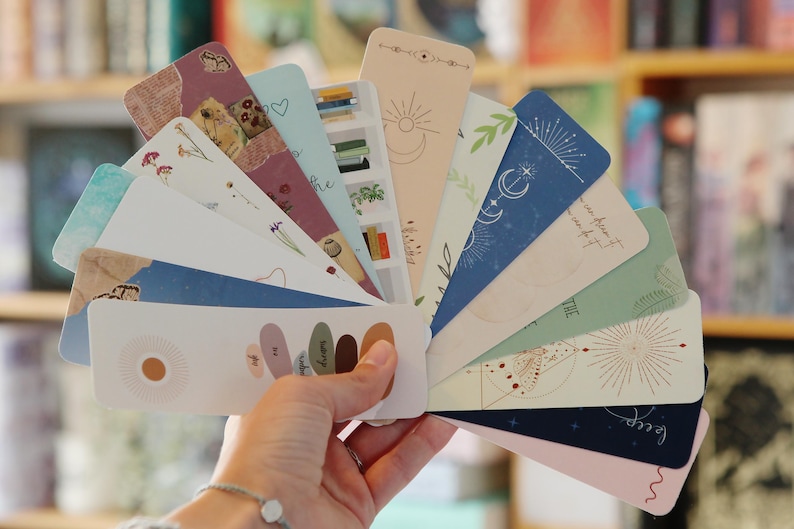 May include: A set of 12 colorful bookmarks with various designs, including floral patterns, celestial motifs, and abstract shapes. The bookmarks are arranged in a fan shape and feature a variety of colors, including pink, blue, green, and brown.