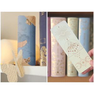 May include: Two bookmarks with different designs. The first bookmark is blue with a butterfly and a piece of paper with text. The second bookmark is light green with a geometric design featuring a butterfly, moons, and stars.