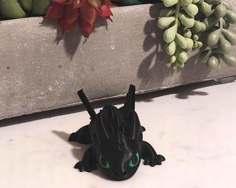 Puede incluir: Figura negra de un dragón con ojos verdes. El dragón está tumbado con las alas y las orejas levantadas. Sobre una superficie blanca, un macetero de hormigón con suculentas artificiales en el fondo.