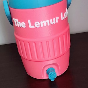 May include: A pink and teal water cooler with the text "The Lemur Lab" in white. The cooler has a spigot and two handles. The cooler is on a dark brown surface.