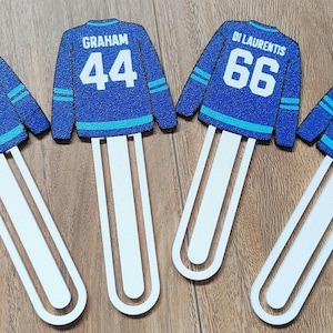 Op de afbeelding: Vier boekenleggers in de vorm van blauwe en groenblauwe sportshirts met witte accenten. Elk heeft een unieke naam en nummer: TUCKER 46, GRAHAM 44, DI LAURENTIS 66 en LOGAN 22. De shirts zijn bevestigd aan lange, witte, afgeronde stelen.