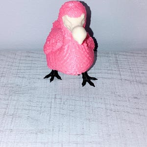 Pode incluir: Estatueta de pássaro rosa com superfície texturizada. O pássaro tem bico branco e pés pretos. A estatueta está sobre uma superfície clara.