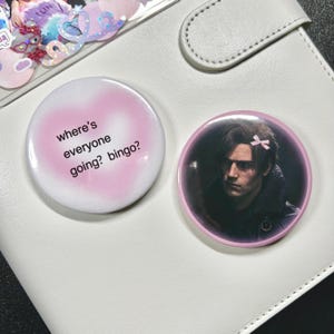 Op de afbeelding: Twee ronde knopen met roze randen. Eén knop toont de tekst "where's everyone going? bingo?" in het wit. De andere knop toont een man met een roze strik. Beide knopen liggen op een witte ondergrond.