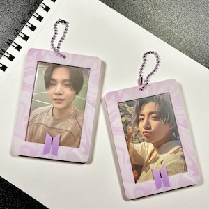 May include: Two rectangular photo holders with a light purple marble design. Each holder has a photo of a person. The holders have a purple BTS logo and a chain for hanging.