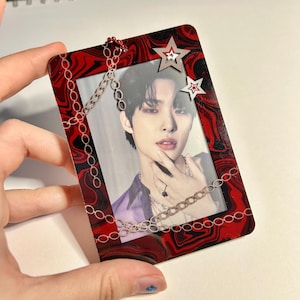 Puede incluir: Una tarjeta fotográfica rectangular con un retrato. La tarjeta tiene un borde rojo y negro veteado, una cadena plateada y dos adornos en forma de estrella. Una cadena de cuentas rojas está unida en la parte superior.