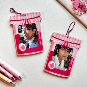 May include: Two pink acrylic keychains shaped like jam jars, each featuring a photo of a person. The keychains have a gingham pattern on the lid and a strawberry graphic. Each keychain has a pink beaded chain.