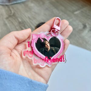Könnte beinhalten: Ein klarer Acryl-Schlüsselanhänger mit einem rosa herzförmigen Rand und einem Foto eines Mannes. Der Schlüsselanhänger hat eine rosa Schleife oben und die Worte "I my boyfriend!" in Rosa. Ein rosafarbener Metallverschluss ist angebracht.