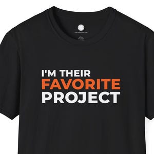 Puede incluir: Camiseta negra de cuello redondo con el texto "I'M THEIR FAVORITE PROJECT" en letras blancas y naranjas. La camiseta es de color negro liso y parece estar hecha de un material suave.