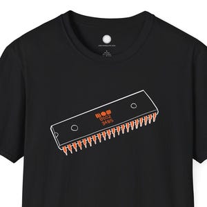 Puede incluir: Camiseta negra con un gráfico blanco y naranja de un chip de computadora. El chip tiene el texto "MOP 3495". La camiseta es de cuello redondo y manga corta.