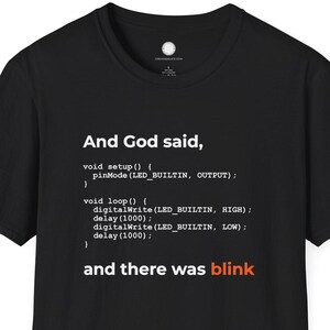 Peut inclure: T-shirt noir avec texte blanc : "And God said, ... and there was blink", présenté comme du code. Le t-shirt est à col rond et à manches courtes.