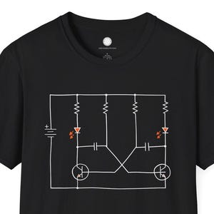 May include: Black t-shirt featuring a white and orange schematic diagram. The diagram includes symbols for resistors, diodes, transistors, and a battery. The design is centered on the front of the shirt.