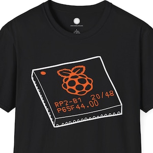 May include: Black t-shirt featuring an orange graphic of a computer chip with a raspberry logo. The chip design includes the text "RP2-B1 P65F44.00 20/48". The shirt is a short-sleeved crew neck.