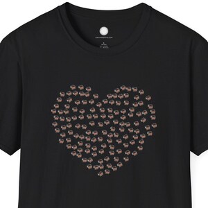 Pode incluir: Camiseta preta com um design em forma de coração feito de pequenas formas castanhas claras. A camiseta tem mangas curtas e gola redonda. O design está centrado na frente.