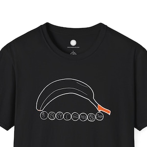May include: Black t-shirt featuring a white outline of a banana with an orange stem. Below the banana are seven small circles with text. The shirt is a classic crew neck style.