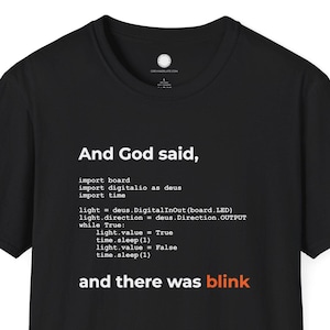 Może przedstawiać: Czarny t-shirt z białym napisem "And God said, ... and there was blink". Tekst przypomina kod. Koszulka ma okrągły dekolt i krótkie rękawy.