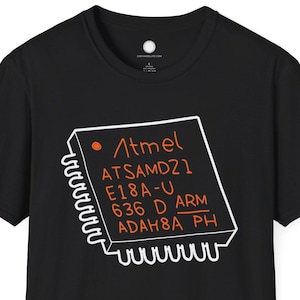 以下が含まれることがあります： 「Atmel ATSAMD21 E18A-U 636 D ARM ADAH8A PH」の文字が入った、白とオレンジのグラフィックが特徴の黒いTシャツ。クルーネックと半袖のデザインです。
