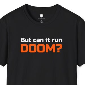 May include: Black t-shirt with the text "But can it run DOOM?" in white and orange. The shirt is a short-sleeved crew neck style, suitable for casual wear. The text is centered on the front of the shirt.