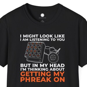 Puede incluir: Camiseta negra con texto y gráficos en blanco y naranja. El texto dice: "I MIGHT LOOK LIKE I AM LISTENING TO YOU BUT IN MY HEAD I'M THINKING ABOUT GETTING MY PHREAK ON." El gráfico incluye un teclado telefónico y un teléfono de marcación rotatoria.