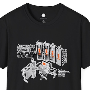 May include: Black t-shirt with a graphic illustration of a new era of integrated electronics. The design includes text and images of computer components and a person working on a computer. The shirt is made of a soft material.