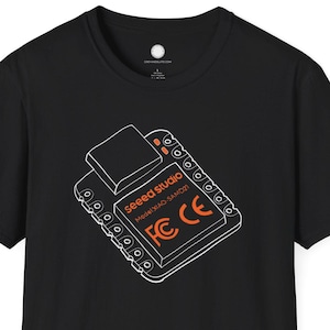 Può includere: T-shirt nera con grafica bianca e arancione di un circuito stampato, con il testo "Seeed Studio" e "FCC CE". La maglietta ha girocollo e maniche corte. Il design è centrato sul davanti.