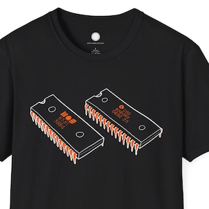 May include: Black t-shirt featuring a graphic of two orange and white computer chips. The chips have the text "NOS" and "6502" printed on them. The shirt is a short-sleeved crew neck.