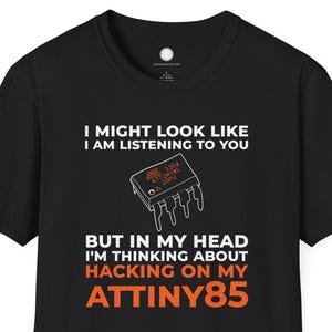 以下が含まれることがあります： 白とオレンジのテキストとマイクロチップのグラフィックが入った黒いTシャツ。「I MIGHT LOOK LIKE I AM LISTENING TO YOU BUT IN MY HEAD I'M THINKING ABOUT HACKING ON MY ATTINY85」と書かれています。