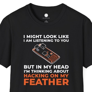 以下が含まれることがあります： 「I MIGHT LOOK LIKE I AM LISTENING TO YOU BUT IN MY HEAD I'M THINKING ABOUT HACKING ON MY FEATHER」の文字が白とオレンジでプリントされた黒いTシャツ。基板のグラフィックが特徴です。