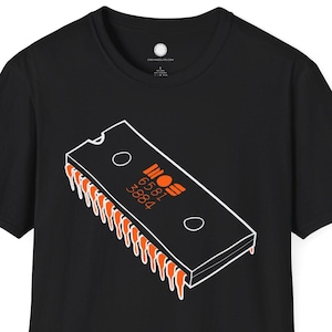 May include: Black t-shirt featuring a graphic of a computer chip with the text "6581 3884" in orange. The chip design is outlined in white, with orange pins extending from the bottom. The shirt is a classic crew neck style.
