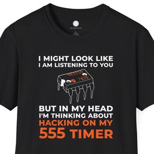 Pode incluir: Camiseta preta com o texto "I MIGHT LOOK LIKE I AM LISTENING TO YOU BUT IN MY HEAD I'M THINKING ABOUT HACKING ON MY 555 TIMER". A camiseta apresenta um gráfico de um circuito integrado 555 timer.