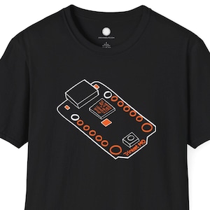 May include: Black t-shirt featuring a white and orange graphic of a circuit board. The design includes a rectangular component, circles, and the text "Trinket M0". The shirt is made of a soft material.