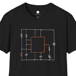 Pode incluir: T-shirt preto com um design de diagrama de circuito branco e laranja. O gráfico inclui resistores, capacitores e um elemento quadrado central. Um design minimalista para entusiastas de tecnologia.