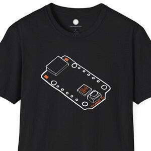以下が含まれることがあります： 白とオレンジ色の回路基板のグラフィックが特徴の黒いTシャツ。デザインには長方形の形、円、オレンジ色の「Trinket」という文字が含まれています。シャツはクルーネックで半袖です。