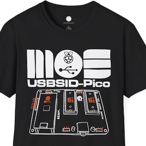 May include: Black t-shirt with a white graphic design. The design includes the text "moe USBSI-Pico" and an illustration of a circuit board with orange and white details. The shirt is made of a soft material.