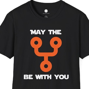 Pode incluir: T-shirt preto com o texto branco "MAY THE BE WITH YOU" e um gráfico laranja estilizado. A t-shirt tem gola redonda.