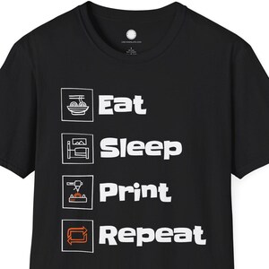 Op de afbeelding: Zwart T-shirt met witte tekst en iconen. De tekst luidt "Eat Sleep Print Repeat" met bijbehorende iconen boven elk woord. Het T-shirt is gemaakt van zacht materiaal en heeft een ronde hals.