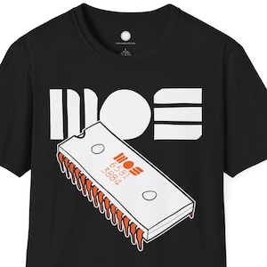 May include: Black t-shirt featuring a white graphic of the word "MOS" and a stylized computer chip with orange accents. The chip has the numbers "6581" and "8584" printed on it. The shirt is a classic crew neck.