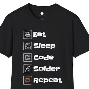 Può includere: T-shirt nera con testo e icone bianche. Il testo recita "Eat, Sleep, Code, Solder, Repeat". Le icone rappresentano una ciotola di cibo, un letto, un computer, un saldatore e un circuito stampato. La maglietta è girocollo.