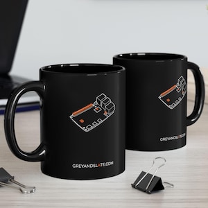May include: Two black coffee mugs with a white and orange circuit board design. The mugs have the text GREYANDSLATE.COM printed on them. The mugs are on a light-colored wooden surface.