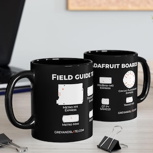 May include: Two black ceramic mugs with white text and diagrams. One mug reads "FIELD GUIDE" and "METRO M4 EXPRESS". The other mug reads "DAFRUIT BOARDS" and "ITSYBITSY MO EXPRESS". The website GREYANDSLATE.COM is also visible.