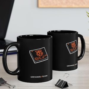 May include: Two black ceramic mugs with a white and orange graphic of a raspberry and text. The mugs are on a light-coloured wooden surface with a laptop, a pencil, and a plant in the background. The website GREYANDSLATE.COM is printed on the mugs.