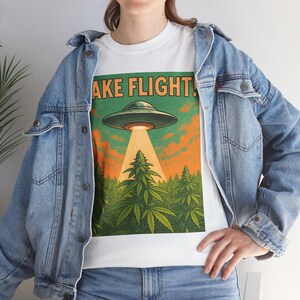 May include: White t-shirt with a graphic of a green UFO beaming a light onto a forest of cannabis plants. The text "TAKE FLIGHT!" is above the UFO. The background features an orange and green sky.