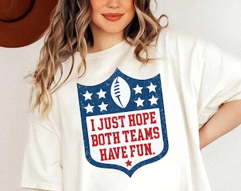 May include: White t-shirt with a blue and red graphic of a football shield with stars and the text "I Just Hope Both Teams Have Fun."